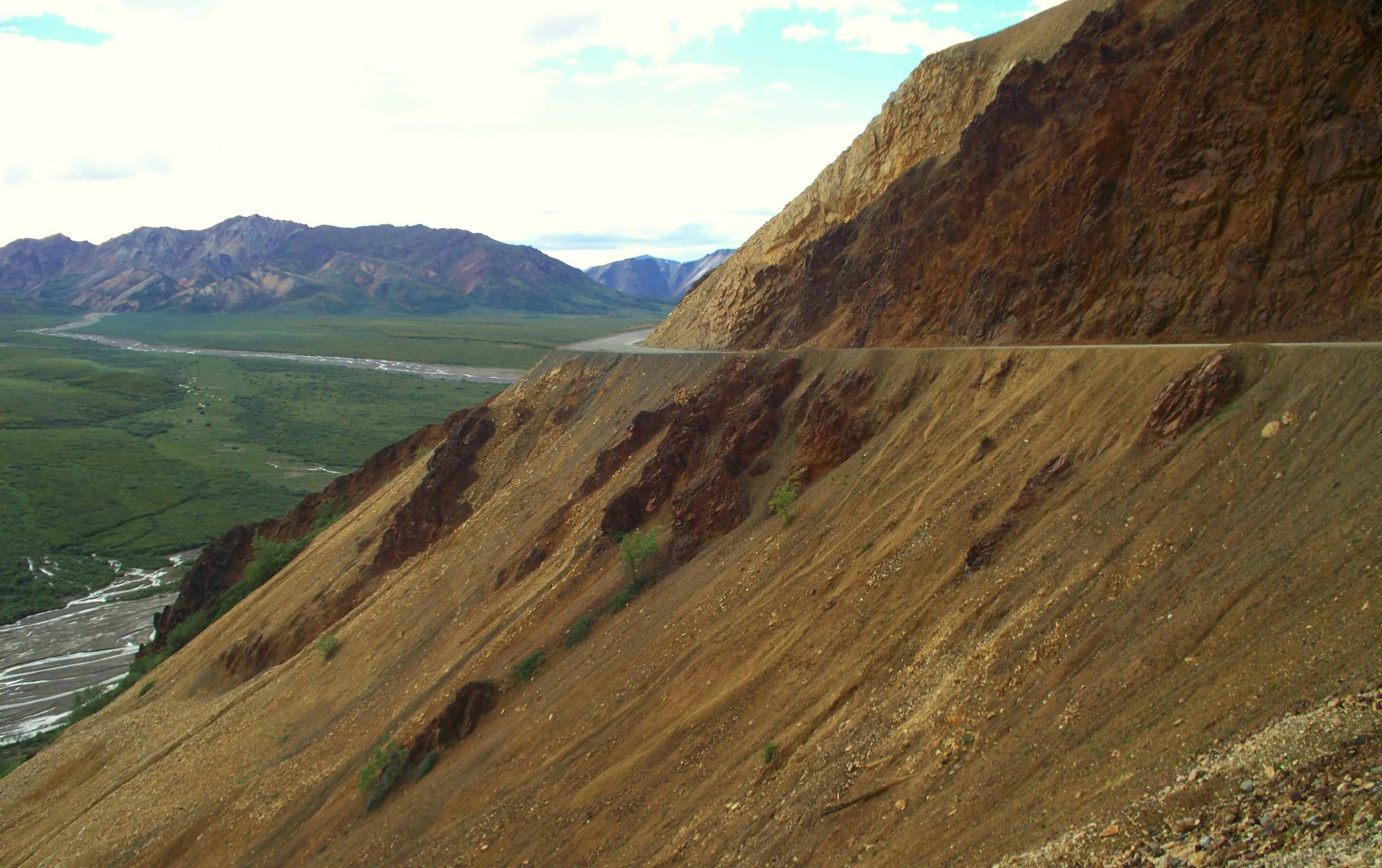 Tan hillside with dusty road cut along it, winding through Alaska's Denali range.