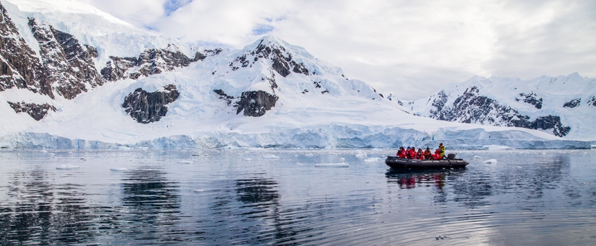 A group wearing red winter parkas sit aboard a black inflatable skiff cruise along the snowy shore line of the Antarctica peninsula.
