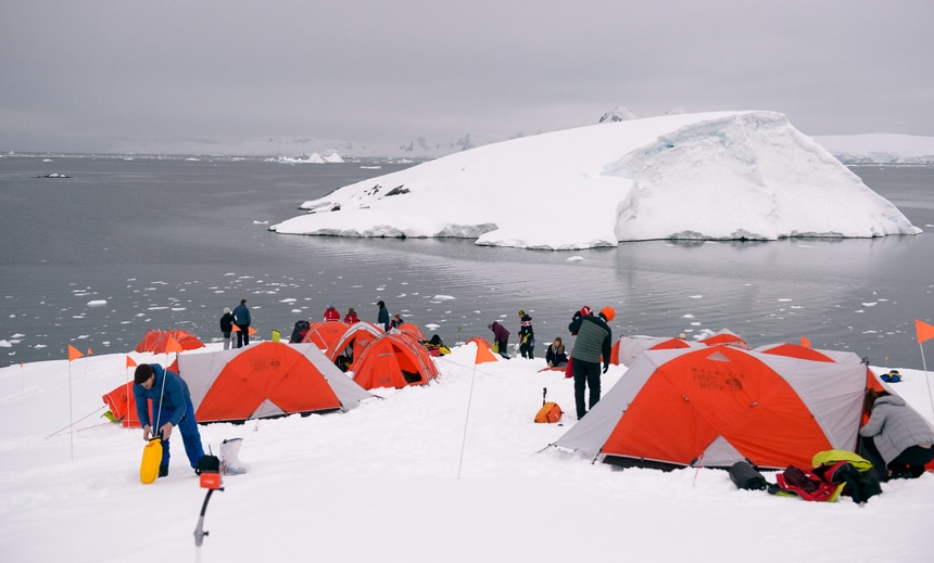 On a grey overcast day cruise guests set up their orange tents on shore in Antarctica for a polar camping activity