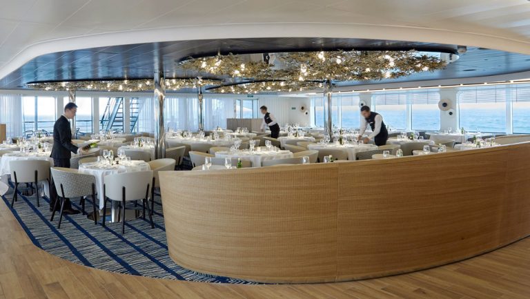 Fancy dining room aboard Le Dumont D'Urville ship, with modern light fixtures, wooden & blue-carpet flooring, white-tablecloth-set tables, plush white chairs & waiters attending to tables.
