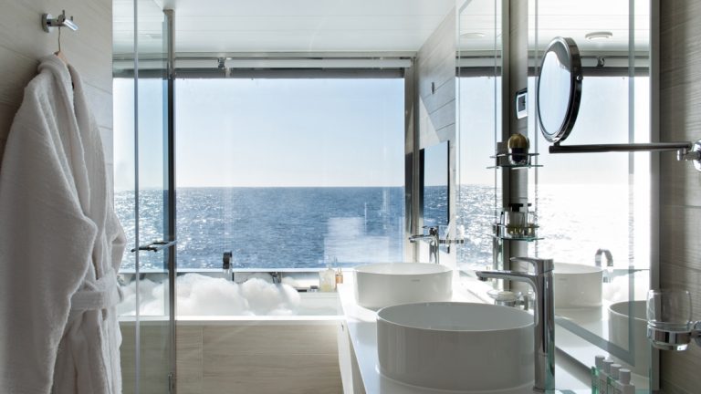 Floor-to-ceiling window beside a large white bathtub with a white robe hanging beside the vanity on Le Jacques Cartier French ship.
