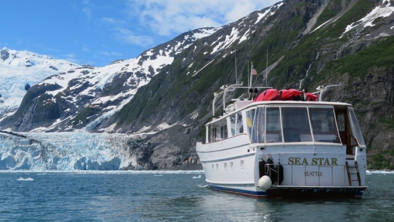Yacht with white exterior & 3 red kayaks on top sits and looks toward a large blue glacier during the Alaska Explorer: Kenai Peninsula & Prince William Sound tour.