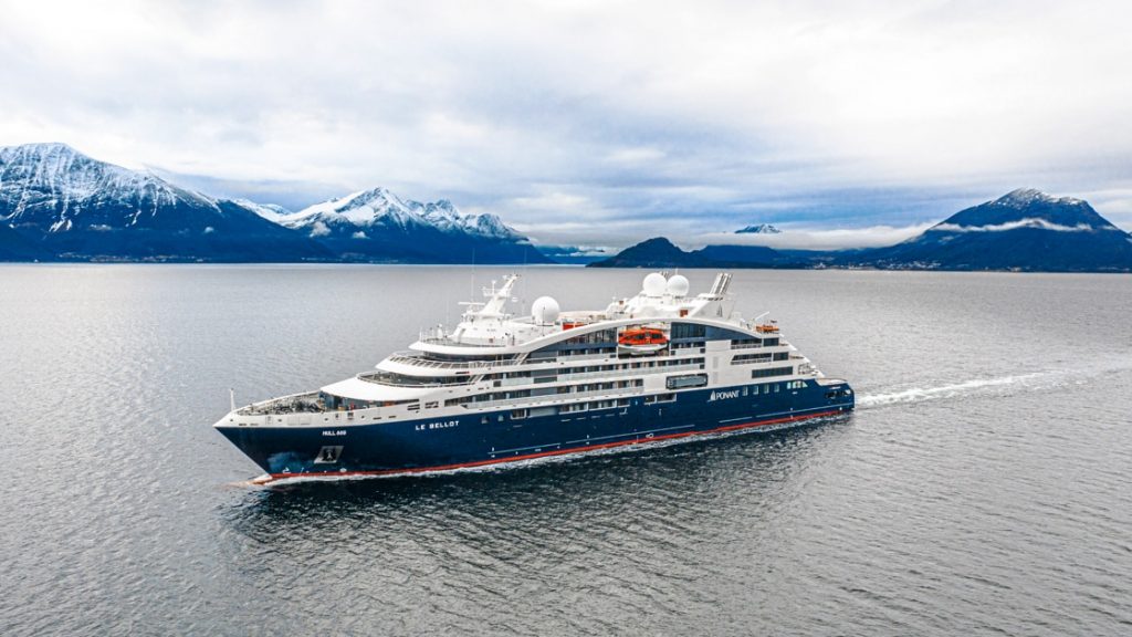 Aerial view of Le Bellot luxury expedition ship with dark blue hull & white upper decks, cruising through calm waters with snow-covered peaks in the background.