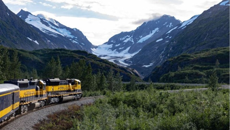 Alaska Railroad train travels through green lowlands amongst snowcapped peaks.
