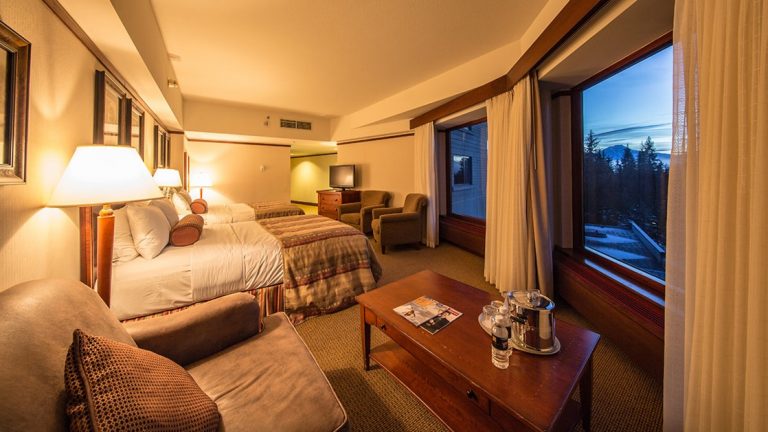 Hotel Alyeska Junior Suite with 2 double beds, padded chairs, TV, wooden coffee table & dresser, beige & tan accents & wraparound windows.