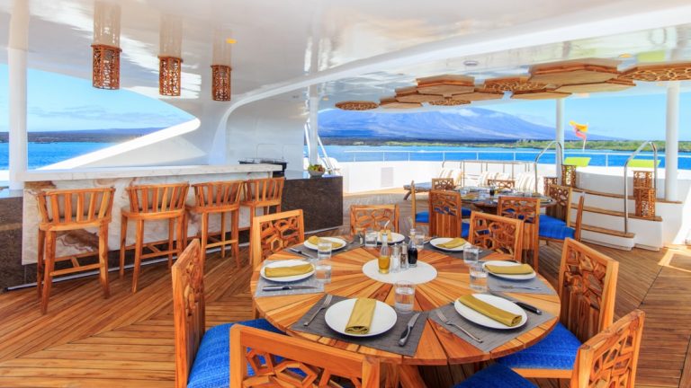 Al fresco dining area set for a meal on Elite catamaran in Galapagos, with teak furniture, marble bar, Jacuzzi & wooden accents.