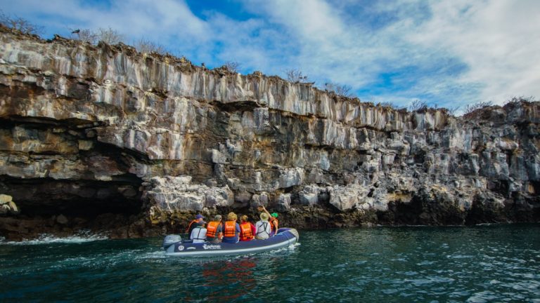Endemic Galapagos catamaran travelers in orange PFDs ride in a Zodiac through calm water along a rocky cliff on a sunny day.