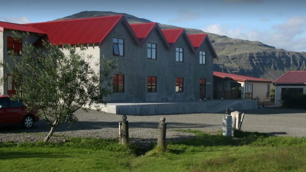 Exterior of Iceland Hotel Smyrlabjorg, with grass, stone building with red metal roof & big windows, small tree & mountains behind.