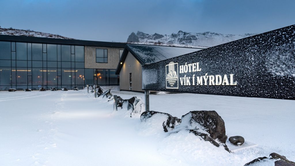 Exterior ground-level view of Iceland Hotel Vik i Myrdal in winter at dusk, with glass walls & illuminated black rectangular sign.
