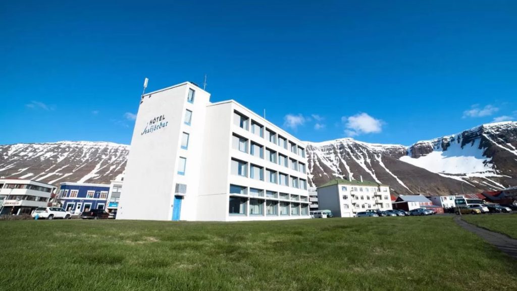 Exterior view of Iceland Hotel Isafjordur, a 4-story white building in the center of town, with green grass & mountains behind.