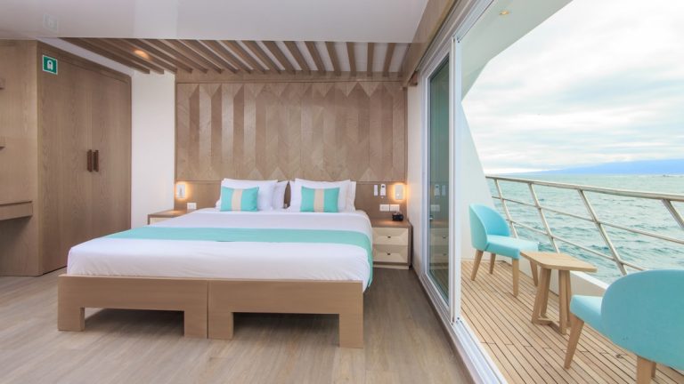 Double bed with white, beige & teal bedding in room with white & wood accents & glass wall with door onto private balcony on M/C Endemic.