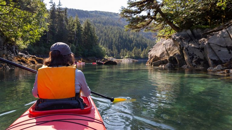 View from behind woman paddling a red kayak in calm waters among rocky tree-lined shore in Alaska.