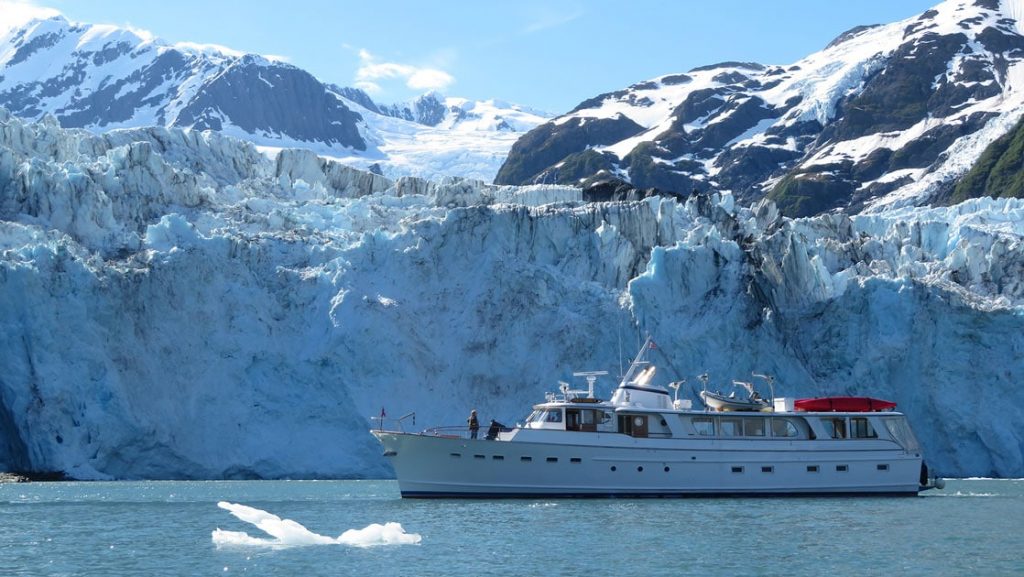 Small white yacht idles in front of a large blue & white glacier on a sunny day in Alaska, with iceberg floating beside the ship.