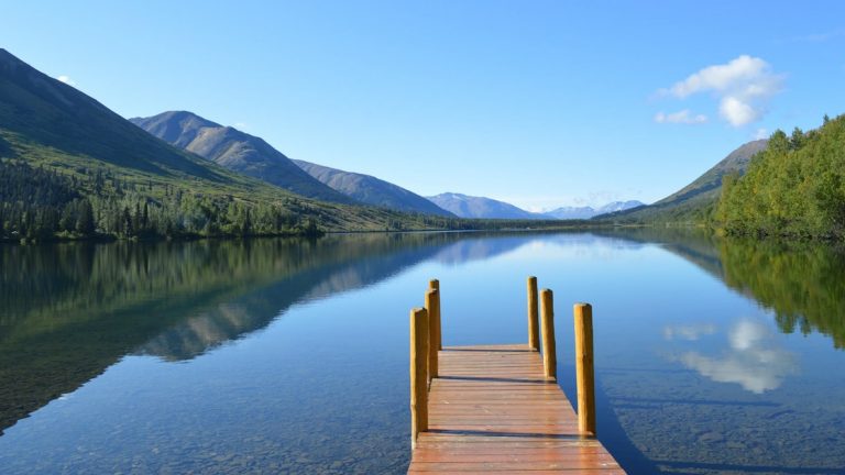 Empty wooden dock leads onto glassy Summit Lake surrounded by green mountains on a clear, sunny day in Moose Pass, Kenai, Alaska.