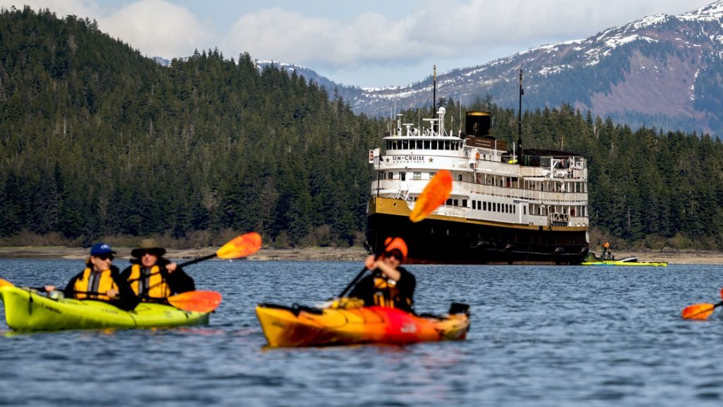 Kayakers in bright lime green and orange kayaks paddlein front of Wilderness Legacy expedition ship with snow-covered mountains in background