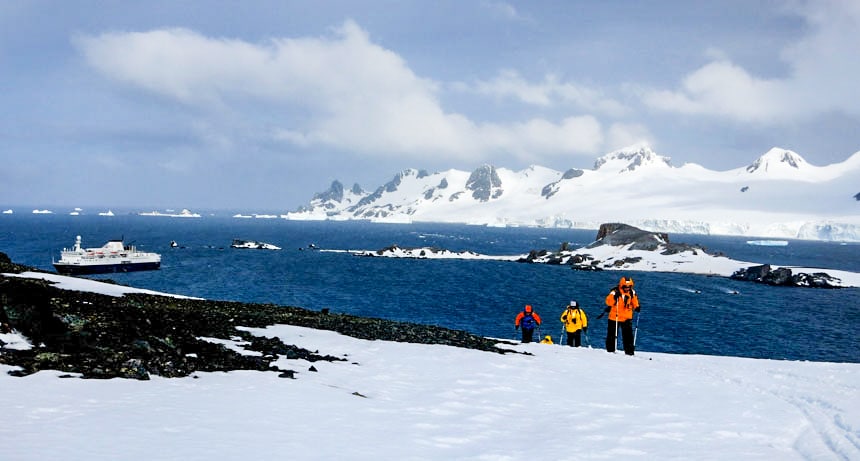 3 viajantes polares em vermelho brilhante, Jaquetas laranja amarelas caminham por um campo de neve em um dia ensolarado. Uma resposta para por que ir para a Antártica.