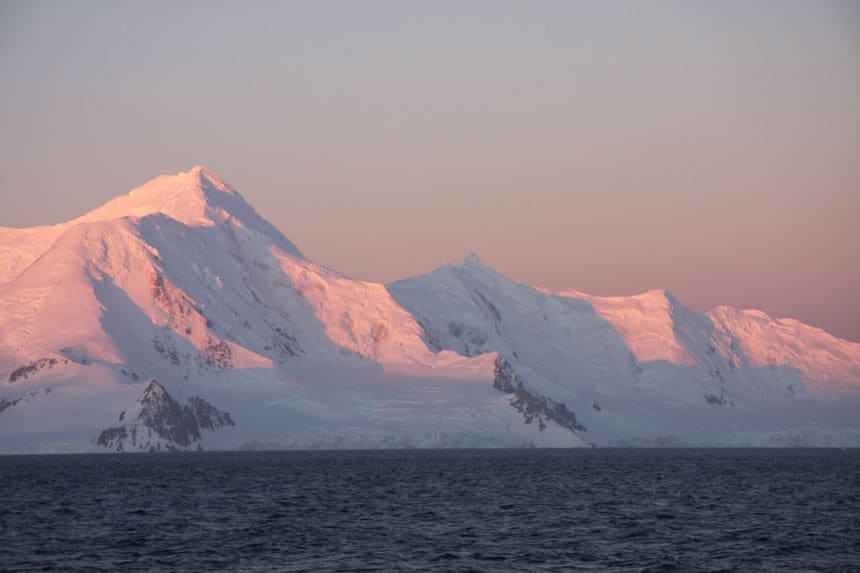 Rosa solnedgang nyanser på snødekte topper ved siden av åpent hav. Et svar for skal jeg gå Til Antarktis?