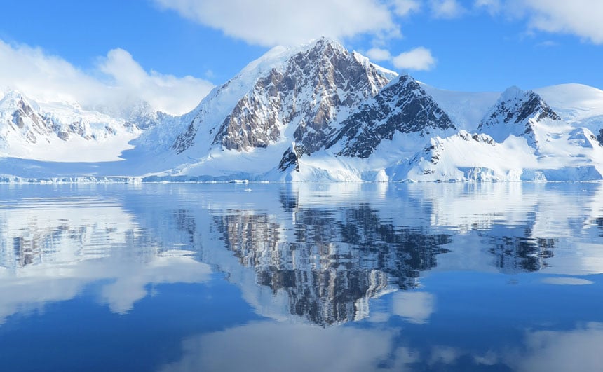 Agua vítrea bajo un cielo azul con algunas nubes blancas un pico rocoso cubierto de nieve. Una razón por la que la gente quiere ir a la Antártida.