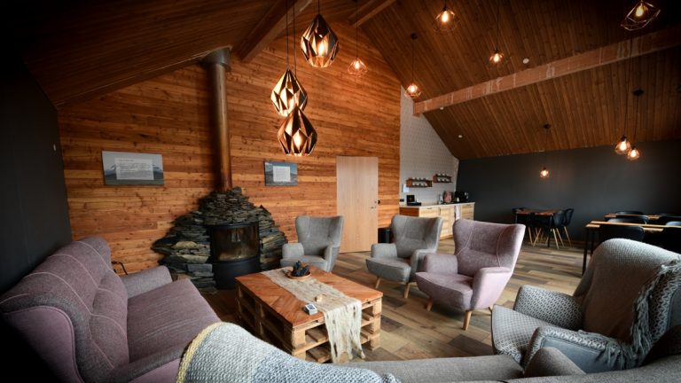 Living room with wooden walls & ceiling, pendant lamps, pink & teal couches & chairs, wood stove, recessed lights & pallet coffee table.