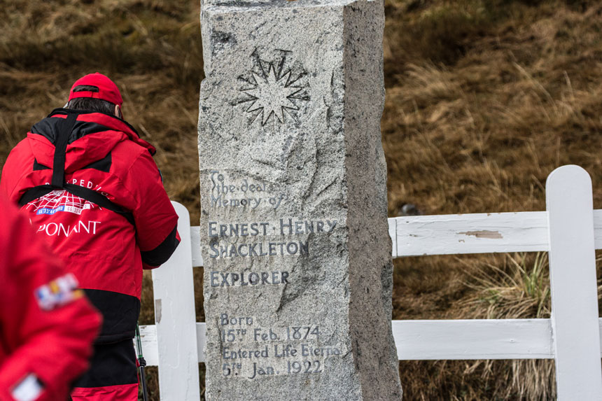 Uomo in giacca rossa si erge davanti alta lapide di granito di Sir Ernest Shackleton. La storia è una risposta al perché andare in Antartide.