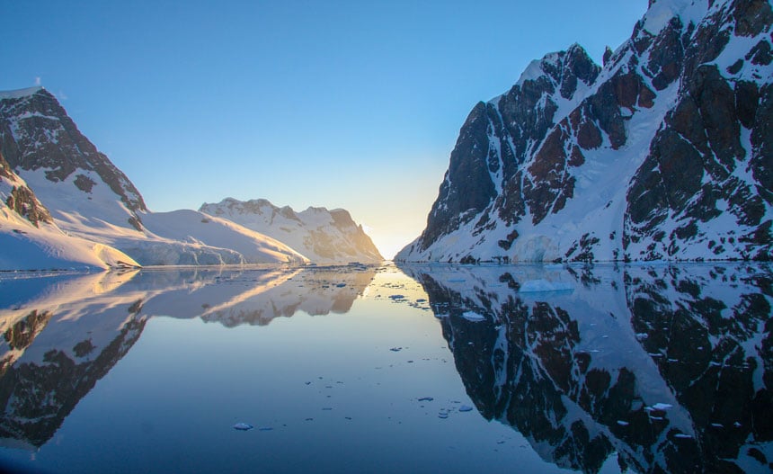 Il sole sorge tra 2 catene montuose rocciose coperte di neve con acqua vetrosa tra di loro. Una risposta a ciò che è così speciale in Antartide.