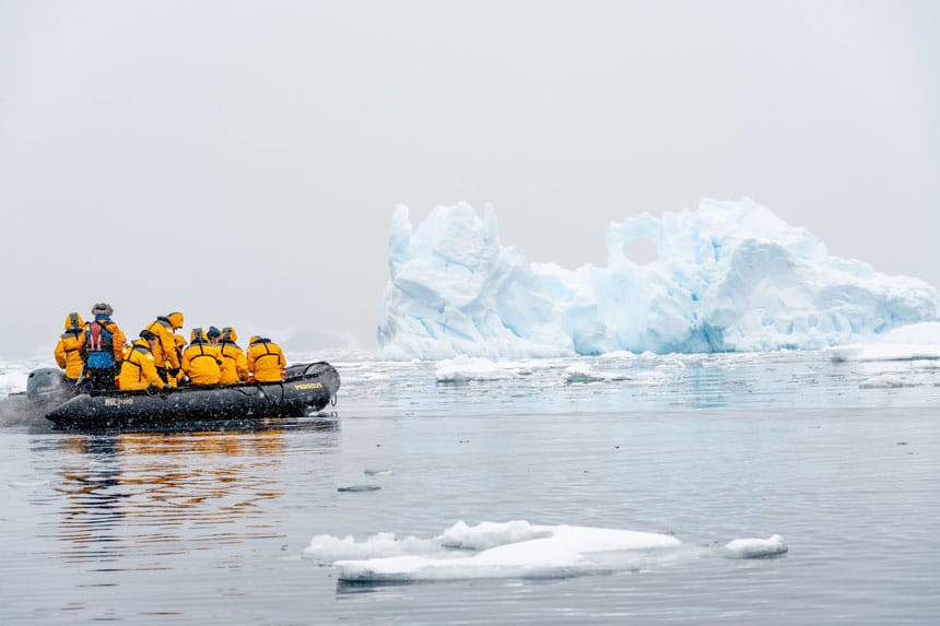 Un grupo de viajeros de la Antártida con chaquetas amarillas se sienta en un barco Zodiac negro mientras navega por un iceberg blanco en un día nevado.