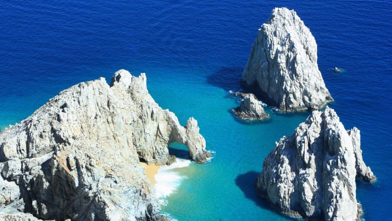 Aerial view of large white basalt rocks rising up out of calm deep blue & turquoise waters on a sunny day in Baja California.