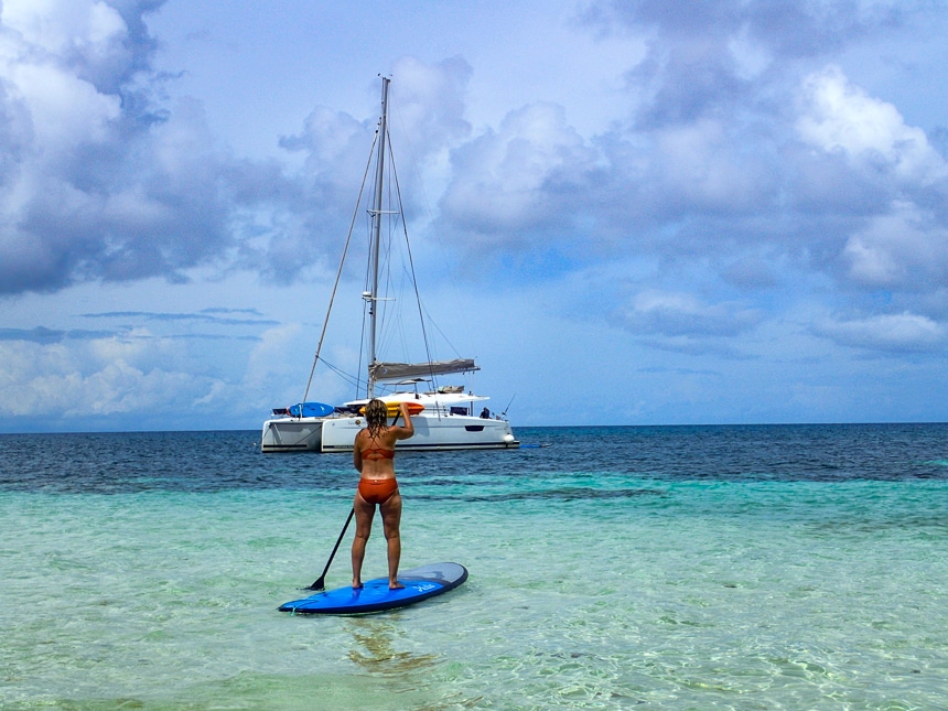 A female in a bathing suit paddles her paddleboard through a crystal clear teal ocean towards a floating white sailboat Belize.