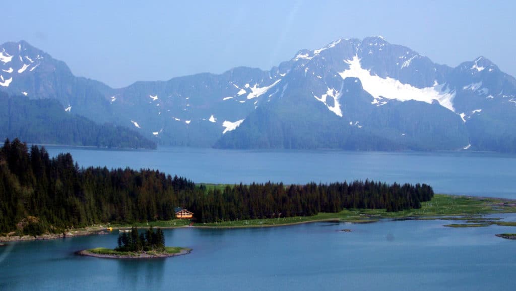 A small coastal cabin on the shore of a peninsula seen from the air with big Alaska mountains behind it