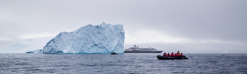 A massive iceberg floats in the dark blue ocean, dwarfing the 6 level expedition ship and inflatable skiff boats around it. 