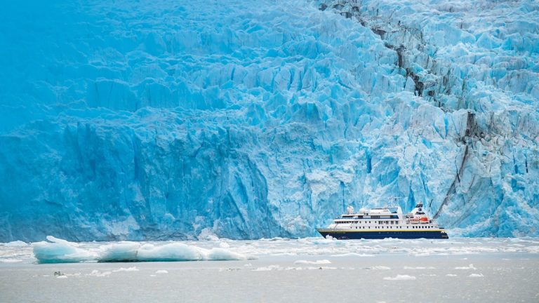Small blue & white expedition ship on a cruise from Chilean fjords to Antarctica, beside a wall of blue ice.