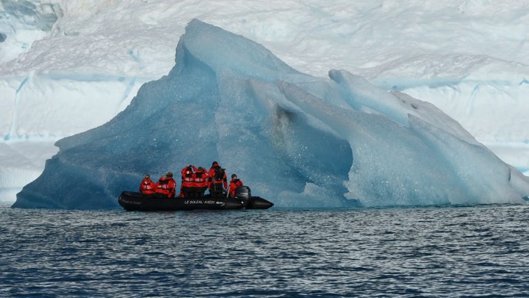 Zodiac with travelers in red jackets cruises by a large iceberg during cruises to Iceland and Greenland aboard Le Commandant Charcot.