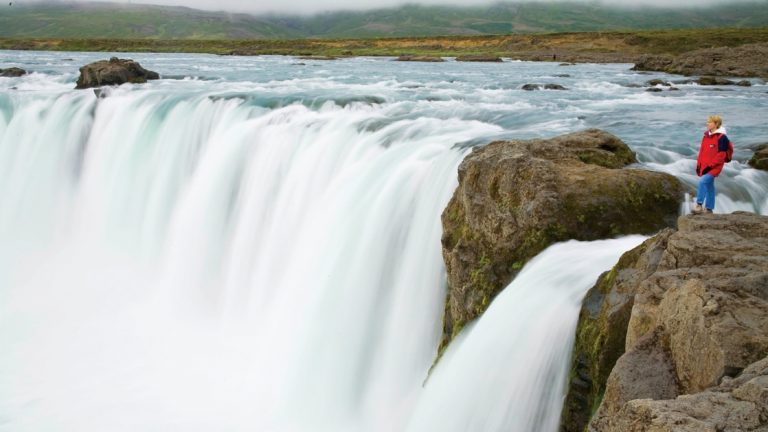 Traveler in a red jacket stands at lip of the giant Godafoss Waterfall of the Gods in Iceland.