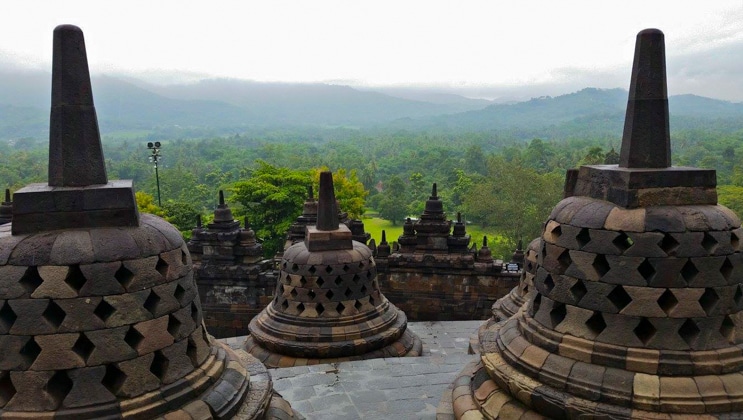 Large stone bell statues with varying shades of tan sit atop a stone terrace looking out over verdant rainforest under bright clouds.