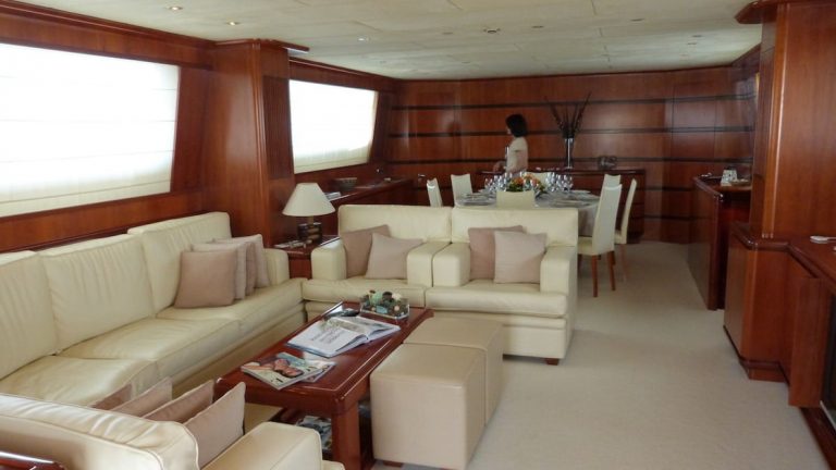 Ship living room beside dining room with wraparound white leather couch, wood coffee table, beige carpet, large windows & wood walls.