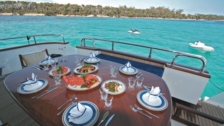 Wood table on outside aft deck of charter yacht, set for a meal, beside turquoise water on a sunny day in Greece.