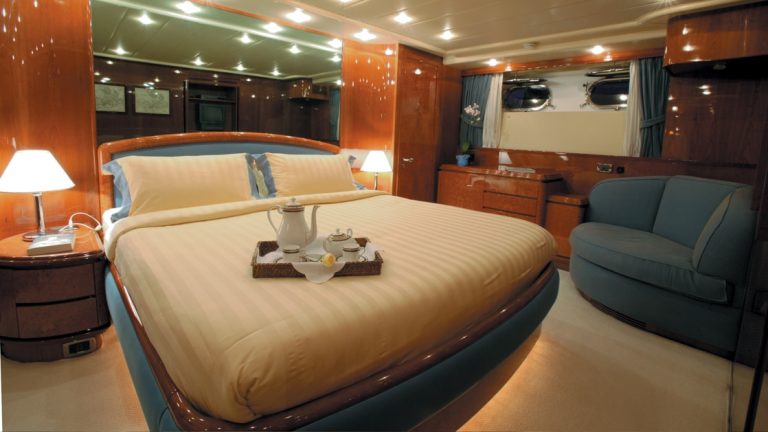 Private yacht cabin with wood veneer, glass & beige accents, blue loveseat, 2 portholes & many recessed ceiling lights.