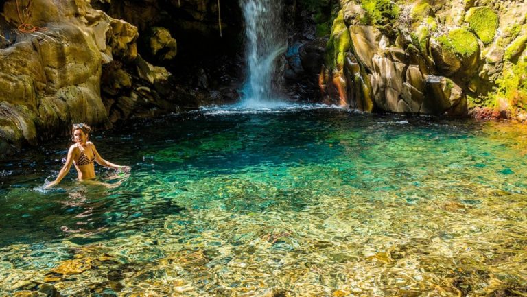A girl in a bikini wades into a clear turquoise pool beneath a small waterfall inside forest of a Costa Rican jungle.