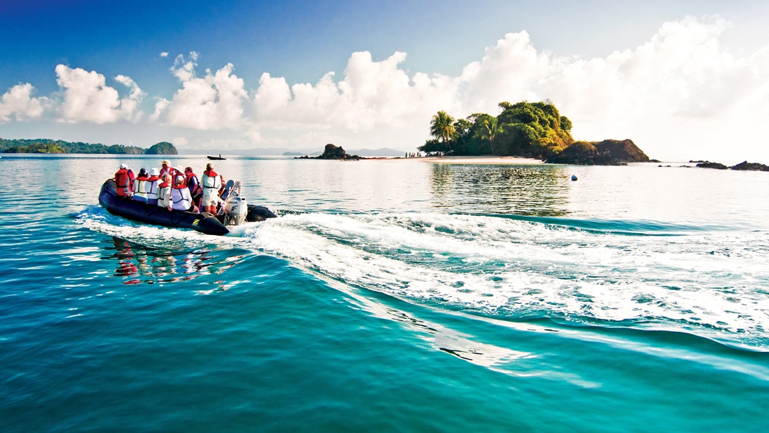 A black inflatable skiff filled with guests in life jackets cruise the teal ocean water by a small island in Costa Rica.