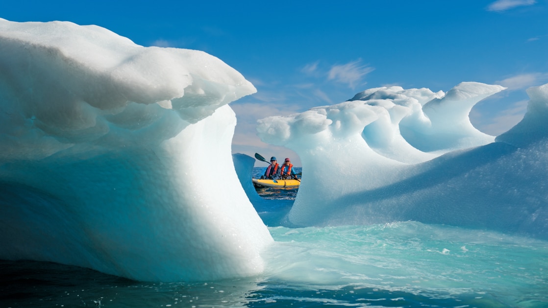 Tandem kayakers in a yellow sit-on-top boat look through an opening between icebergs on a sunny day during the Wild Greenland Escape.