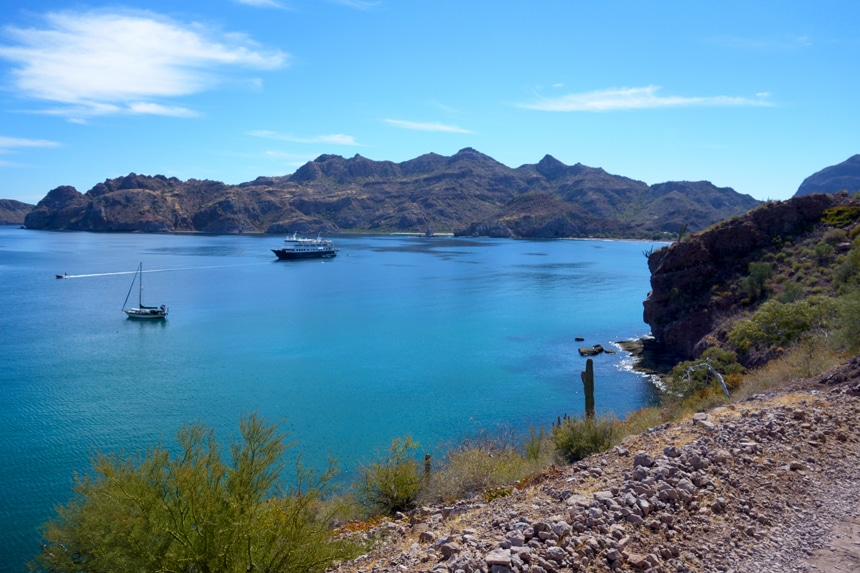 The jagged coastline of the Baja peninsula surrounds the the blue ocean water as a small cruise ship navigates. 