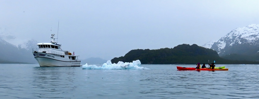 pair of tandem kayakers paddle beside a small iceberg under cloudy skies near the Misty Fjord small ship in Alaska