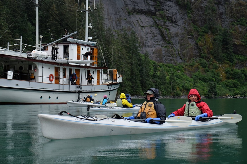 Surrounded by forested granite walls travelers paddle gray double kayaks around the white wooden classic yacht Pacific Catalyst. 