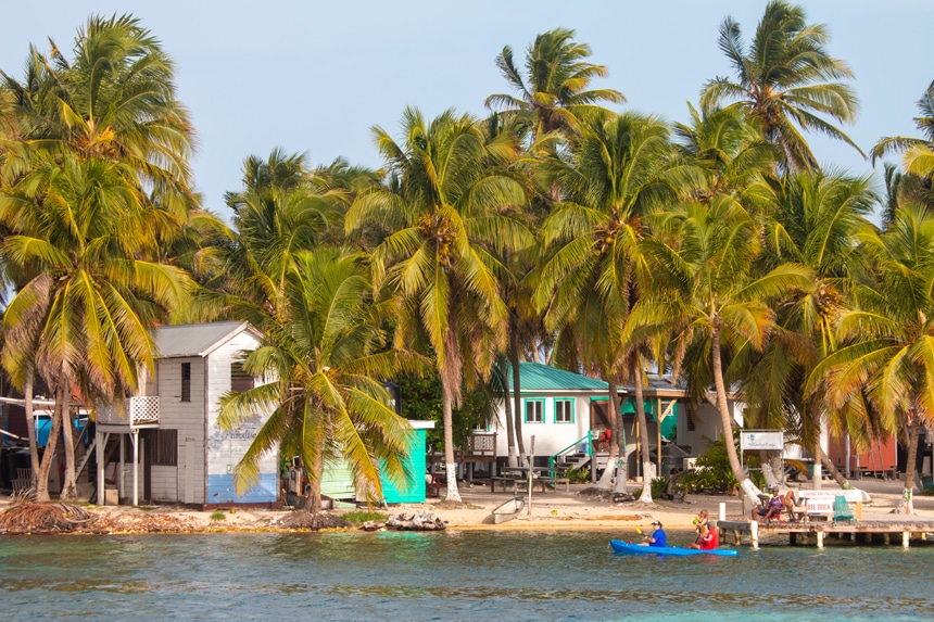 Palm trees and bright colored houses line the shoreline of a sandy island in Belize, people in a double kayak paddle by. 