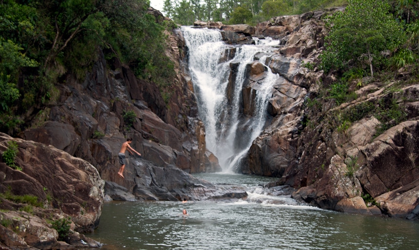 In the Belize jungle, a boy jumps from rocks into a deep pool of water that a large waterfall flows into. 