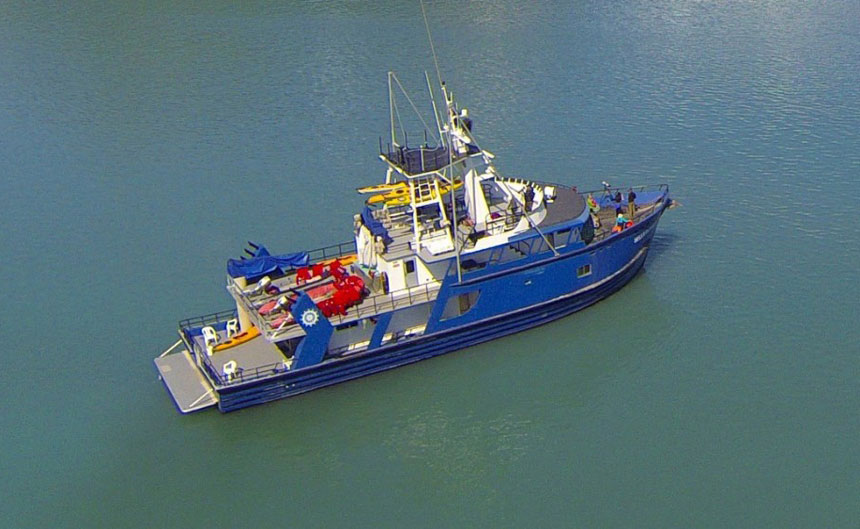 Aerial view of blue Alaska fishing cruise ship Dream Catcher, with 3 passenger decks, swim step, yellow kayaks & lounge chairs.