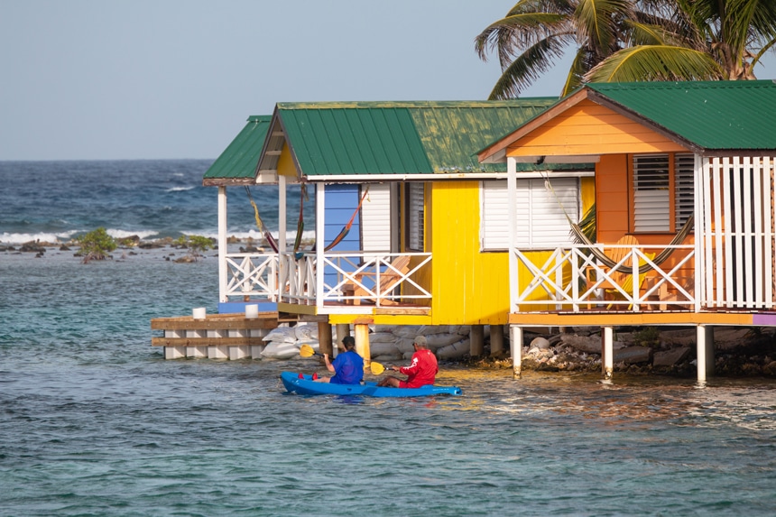 Two kayakers paddle by brightly colored wooden stilt houses that sit over ocean water along a shoreline of a  Belize island.
