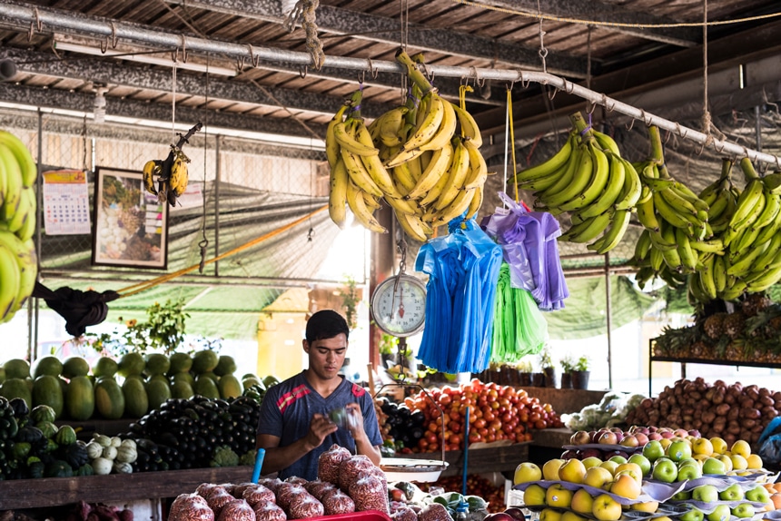 A man stands in between rows of brightly colored fruits and vegetables stands at an outdoor market in Belize. 