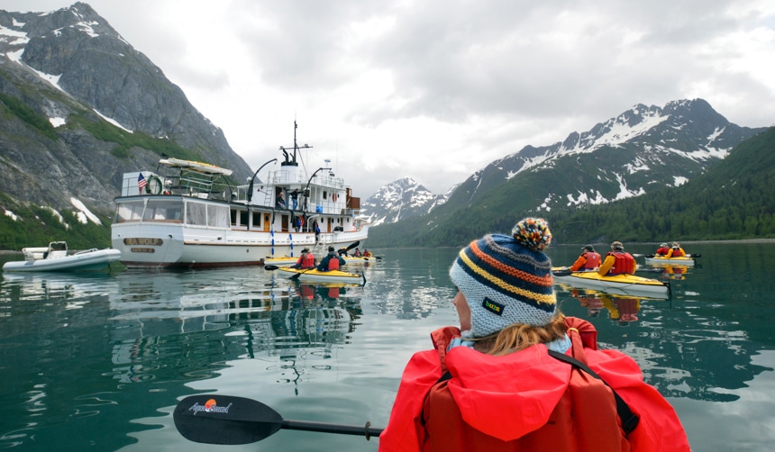 On an overcast day a group of kayakers paddle in a quiet bay in Alaska near their small wooden ship, Sea Wolf. 