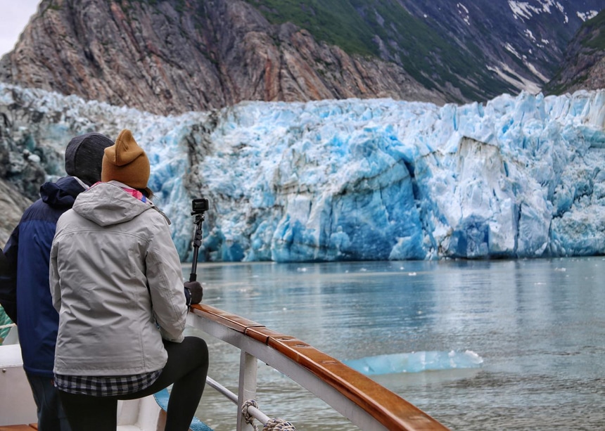 At the bow of a small ship, two travelers lean on the wooden railing as they take in the views of a teal white and blue icy glacier.